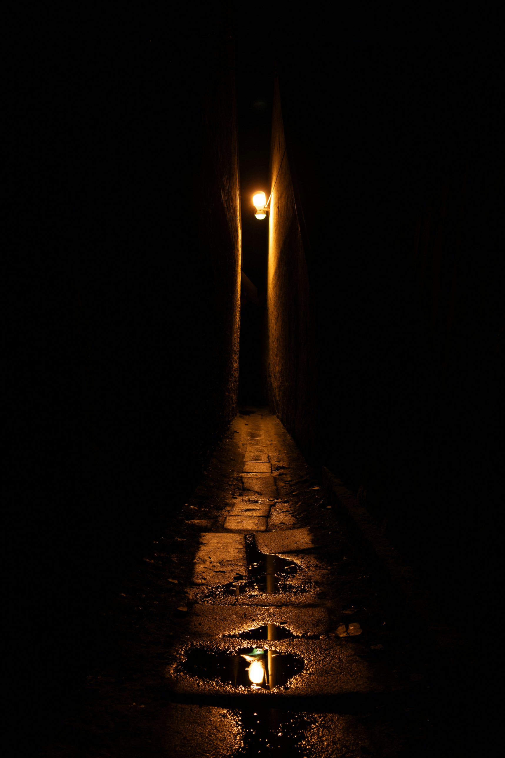 A narrow alley at night, lit by a single lamp, its light reflected in puddles on the wet flagstones.